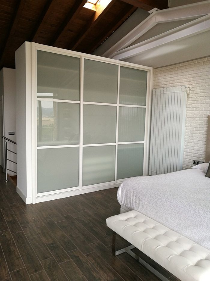 Dormitorio con armario con marco blanco, puertas de vidrio esmerilado y un banco tapizado en blanco.