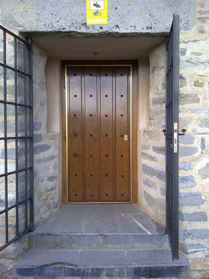 Puerta de madera marrón con tachuelas decorativas y detalles dorados, empotrada en un muro de piedra. Puertas de hierro abiertas a ambos lados.