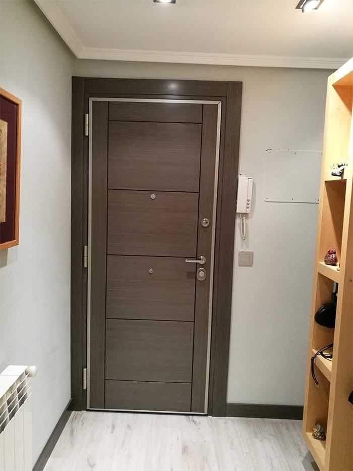 Puerta de entrada de madera gris con detalles plateados en un pasillo con paredes gris claro y un armario de madera.