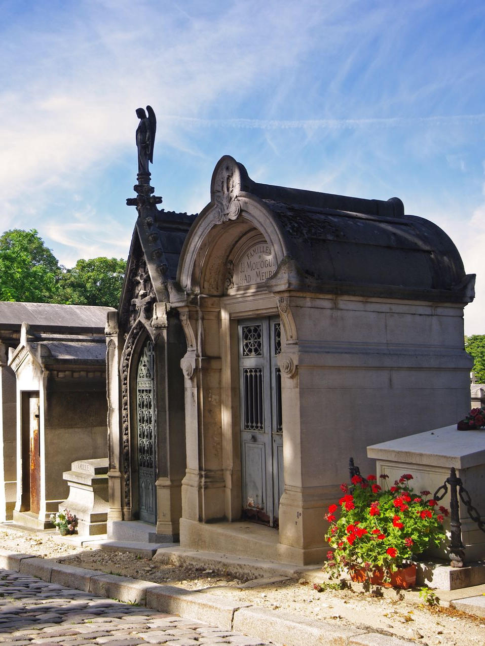 Mausolée en pierre orné d'une statue d'ange et de détails raffinés, avec une petite porte, dans un cimetière. Ciel bleu au-dessus.