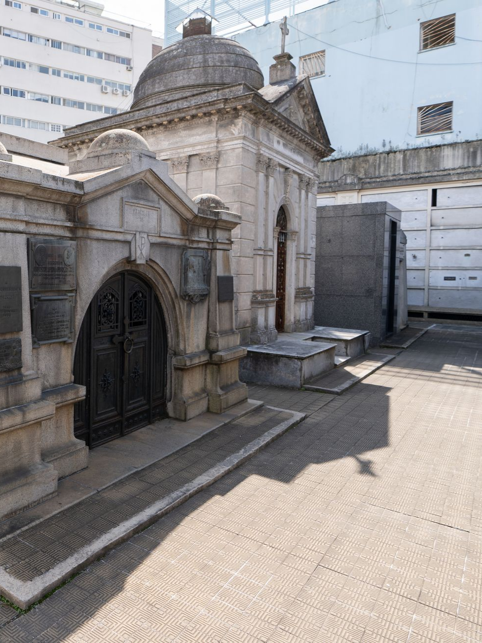 Mausolée en pierre avec entrée voûtée, dôme et petite croix dans un cimetière par une journée ensoleillée.