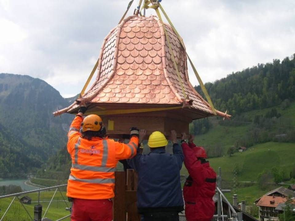 Des ouvriers installent une structure à toit de cuivre, soulevée par une grue, dans un environnement montagneux.