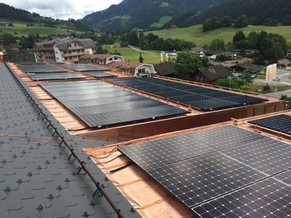 Des panneaux solaires sur le toit d'un bâtiment, avec un paysage montagneux en arrière-plan. On aperçoit des collines verdoyantes et des maisons.