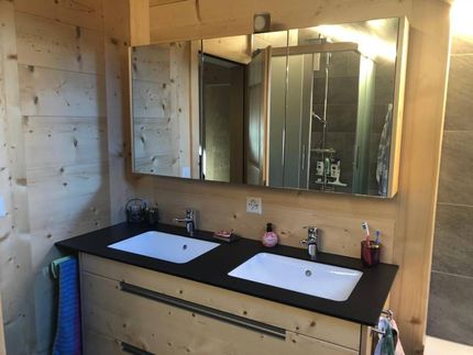 Salle de bains avec double vasque, murs en bois, plan de travail noir, grand miroir et reflet dans la douche.