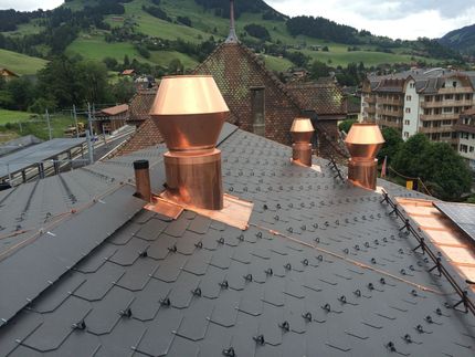 Un toit recouvert de tuiles d'ardoise sombres, avec des aérations et des solins en cuivre, se détachant sur un paysage rural montagneux.