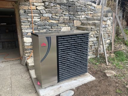 Une pompe à chaleur en métal est installée sur une dalle de béton devant un mur en pierre rustique.
