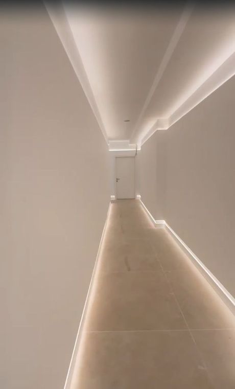 Pasillo minimalista con suelo de baldosas beige, paredes blancas y luces empotradas en el techo que conducen a una puerta cerrada.