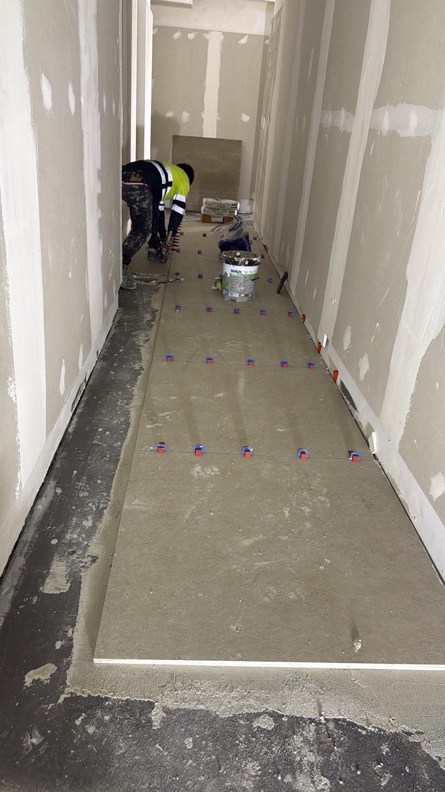 Trabajadores terminando de colocar los paneles de yeso en un pasillo estrecho, con el suelo de hormigón mojado y cubos de pintura.