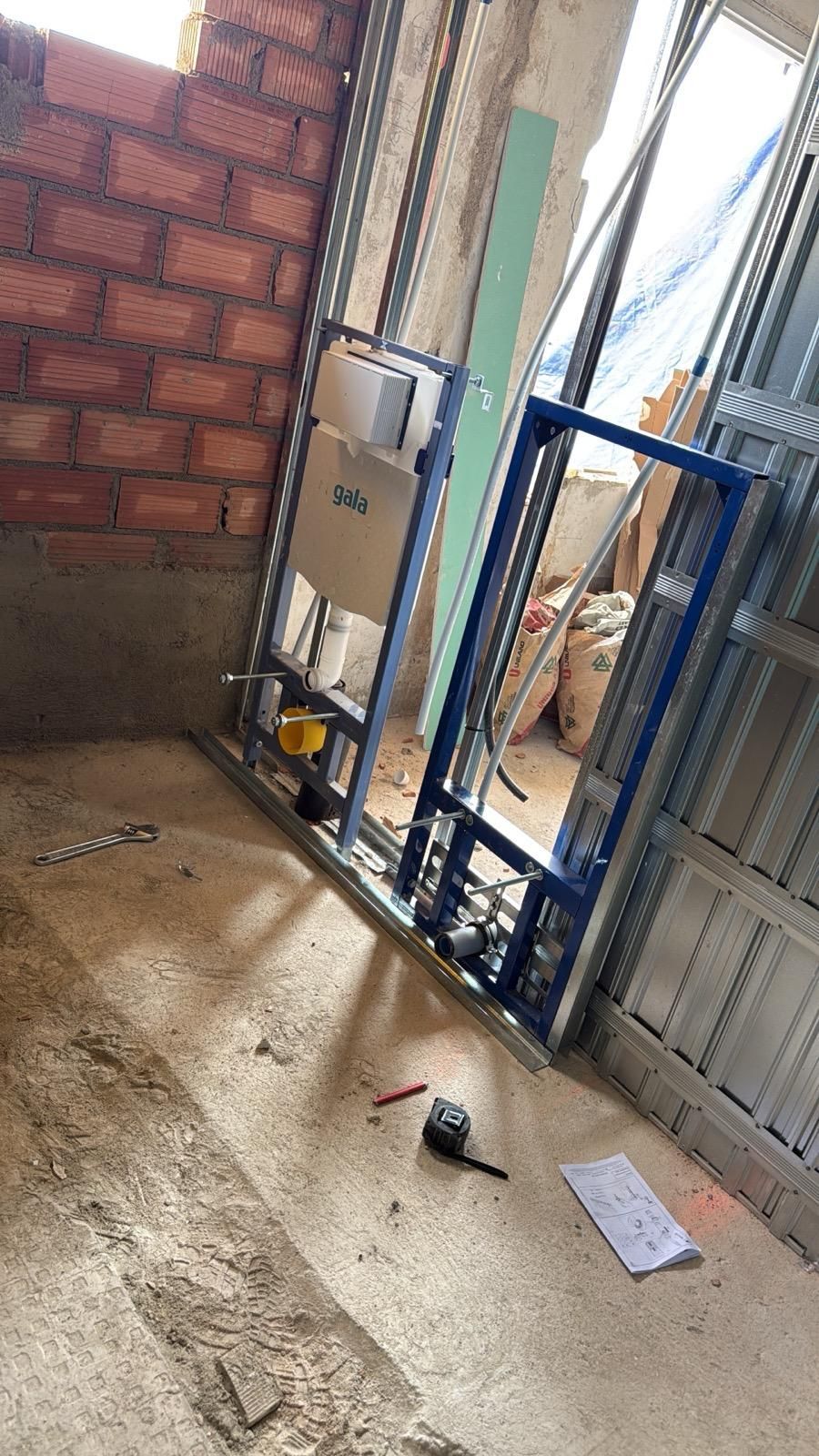 Zona de construcción con una pared de ladrillo, una estructura metálica, herramientas y escombros sobre un suelo de hormigón