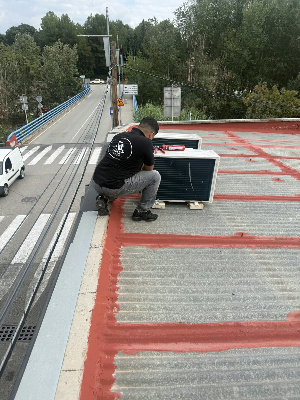 Un trabajador agachado en un tejado, sujetando un maletín negro para equipos cerca de una pasarela de un puente.