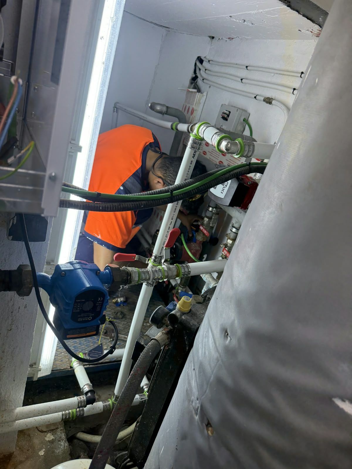 Un técnico con chaleco naranja trabaja en un compartimento de equipos estrecho, rodeado de tuberías y mangueras.