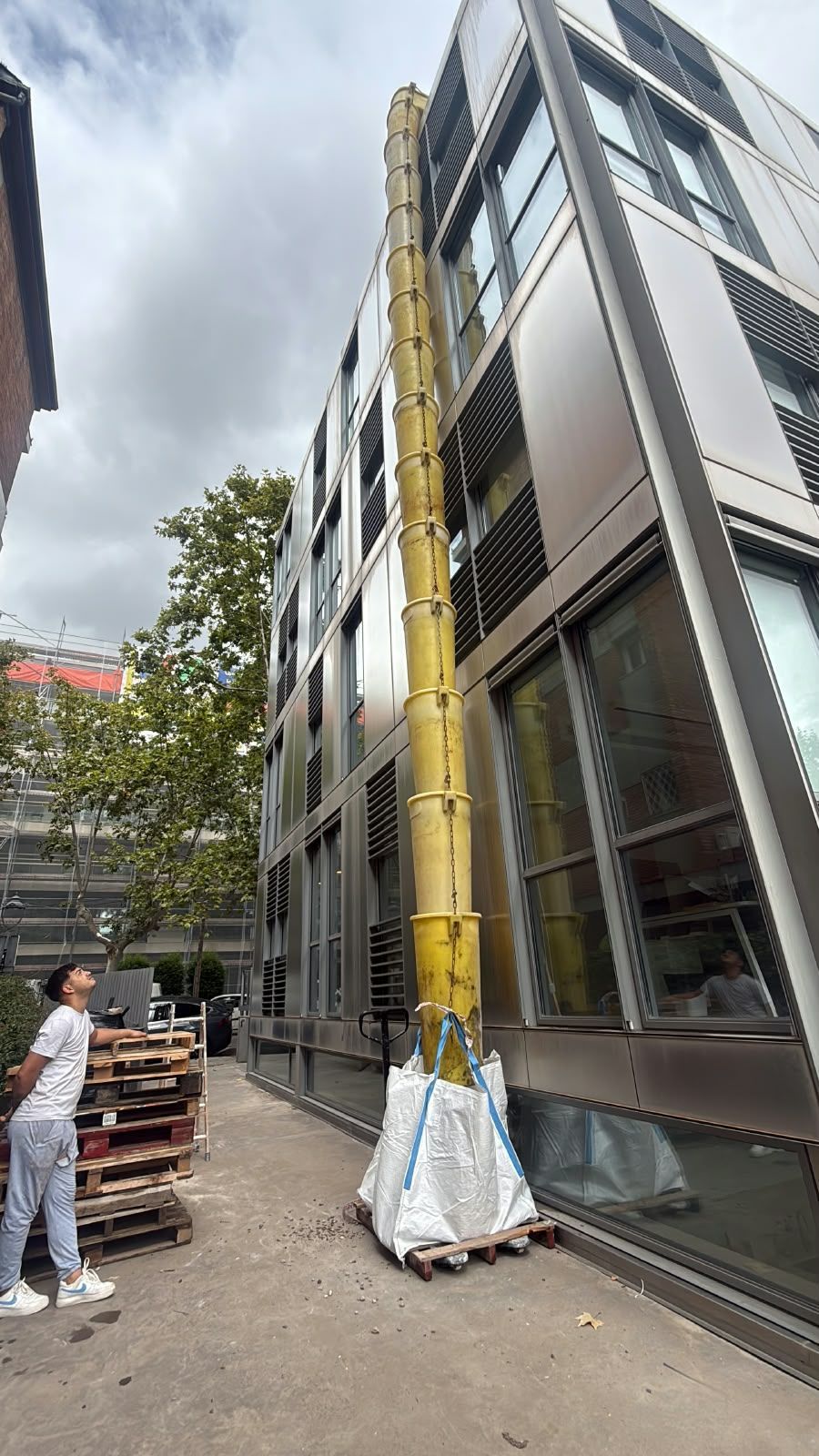 Obrero de la construcción en un andamio junto a un edificio moderno con grandes ventanales y una escalera amarilla.