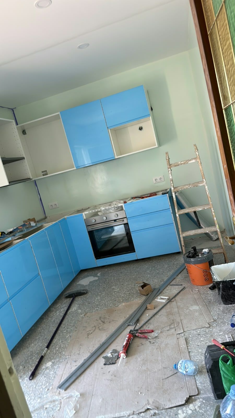 Cocina parcialmente renovada, con armarios azules, escombros en el suelo y una escalera apoyada contra la pared.
