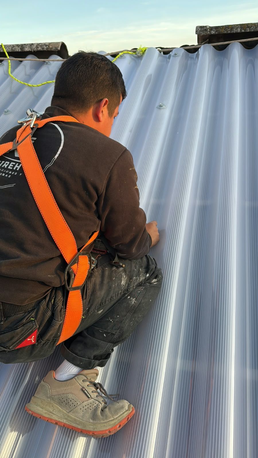 Un trabajador con arnés naranja está arrodillado sobre un tejado de chapa ondulada y utiliza herramientas cerca del borde.