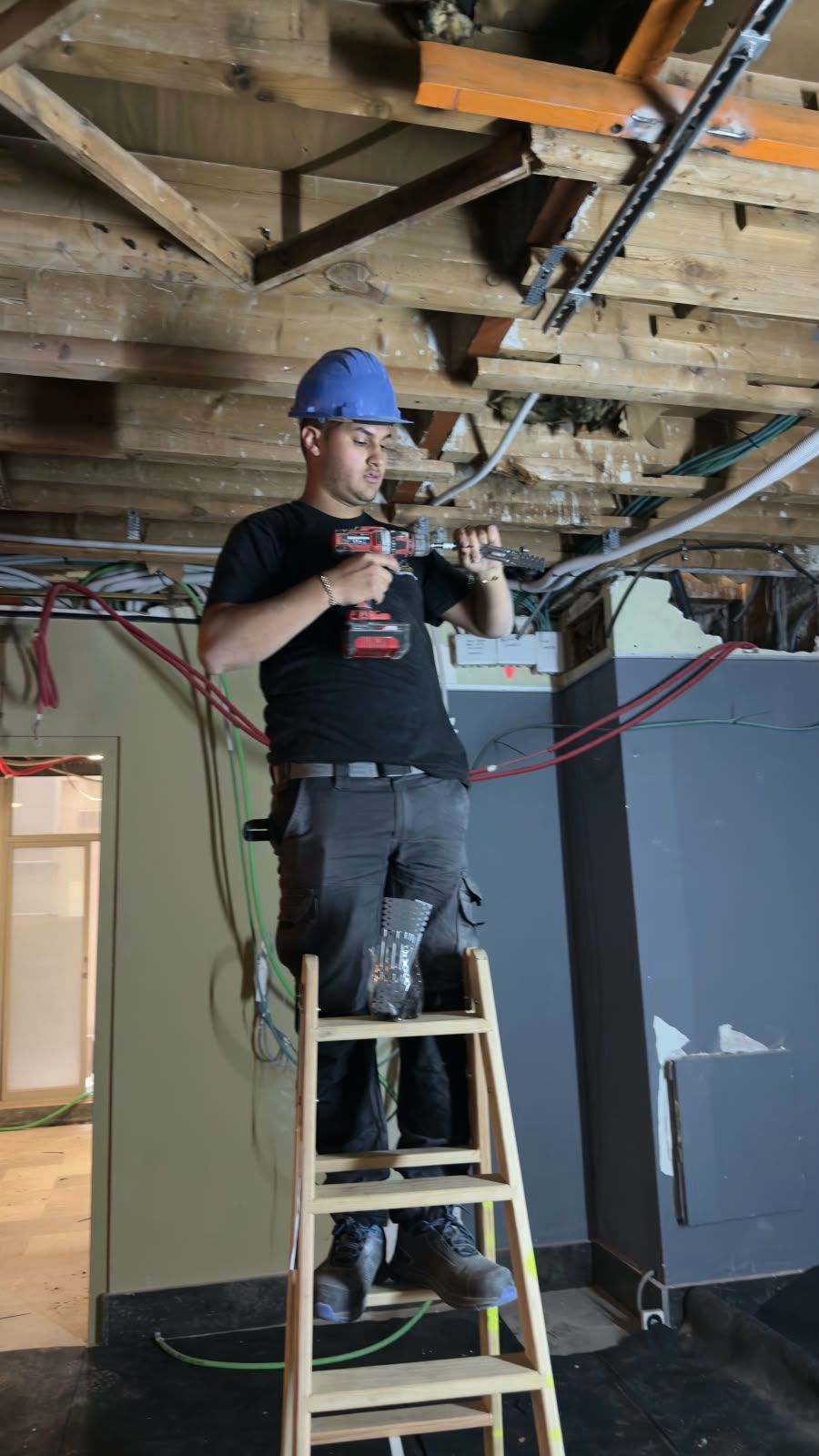 Trabajador subido a una escalera en una habitación sin terminar, ajustando el cableado bajo las vigas del techo a la vista.
