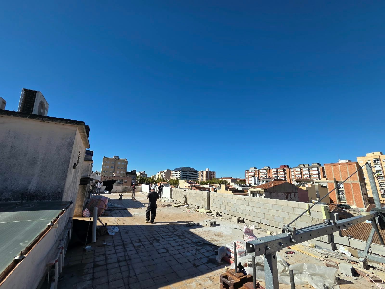 Obra en una azotea con trabajadores, bloques de hormigón y edificios de la ciudad bajo un cielo azul despejado