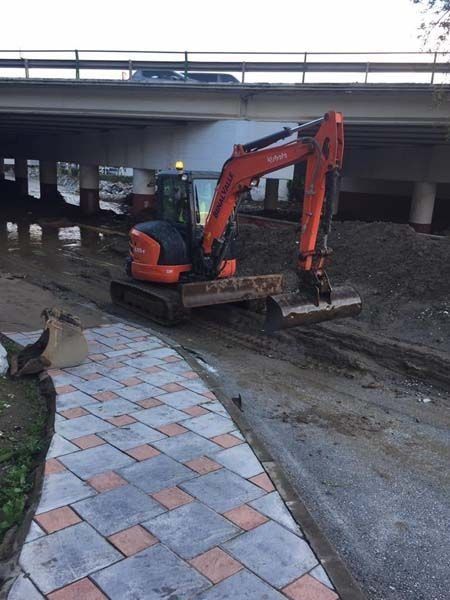 Una pequeña excavadora naranja avanza por un camino de tierra bajo un puente.