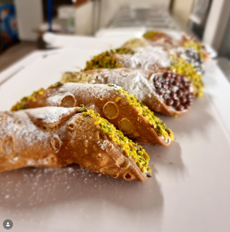 Una fila di cannoli ricoperti di zucchero a velo, disposti su un vassoio bianco e farciti con crema gialla e crema al cioccolato.