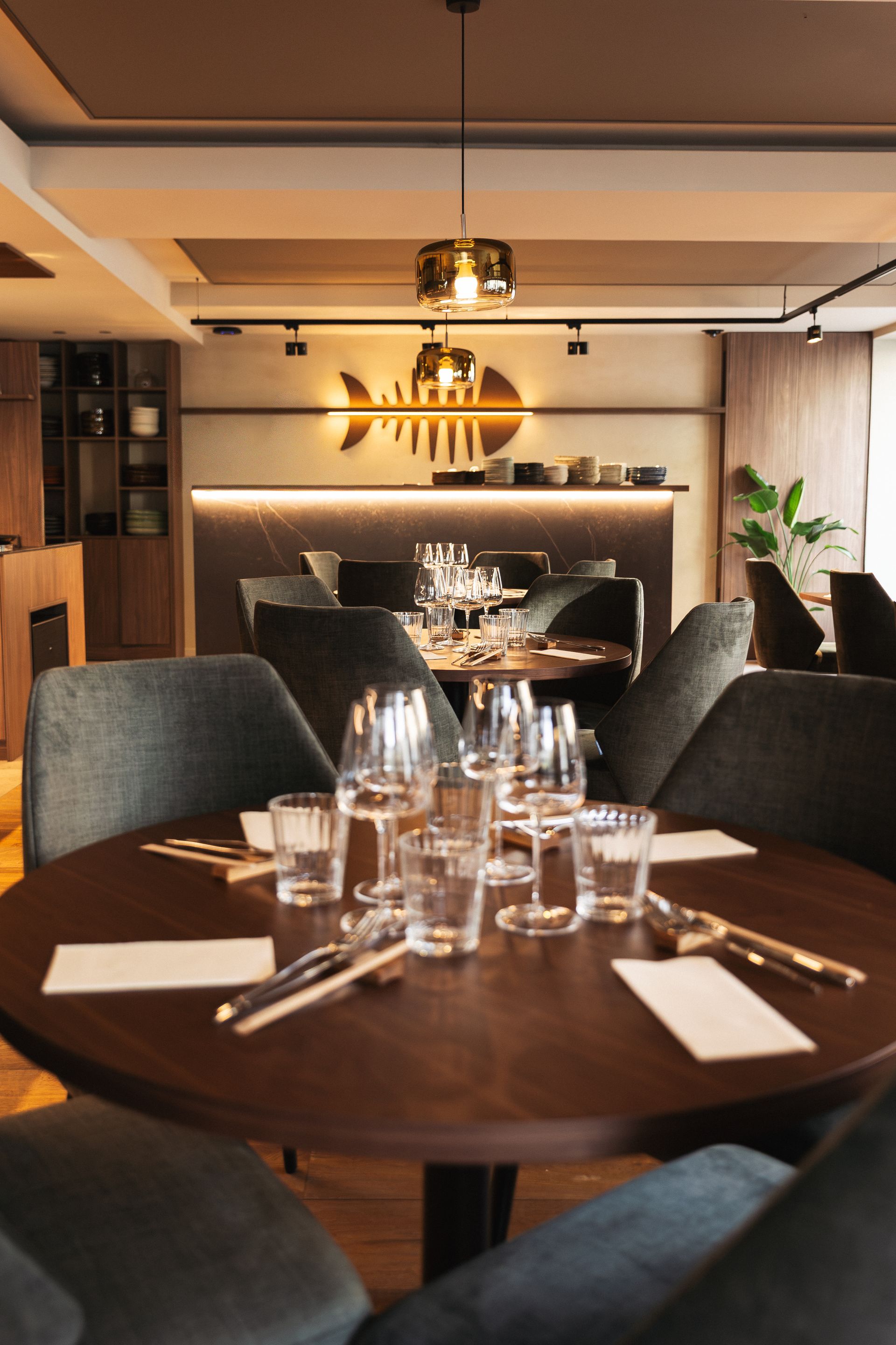Salon moderne avec sièges modulables orange, tables grises, luminaires suspendus et une niche cintrée avec un arbre en pot.
