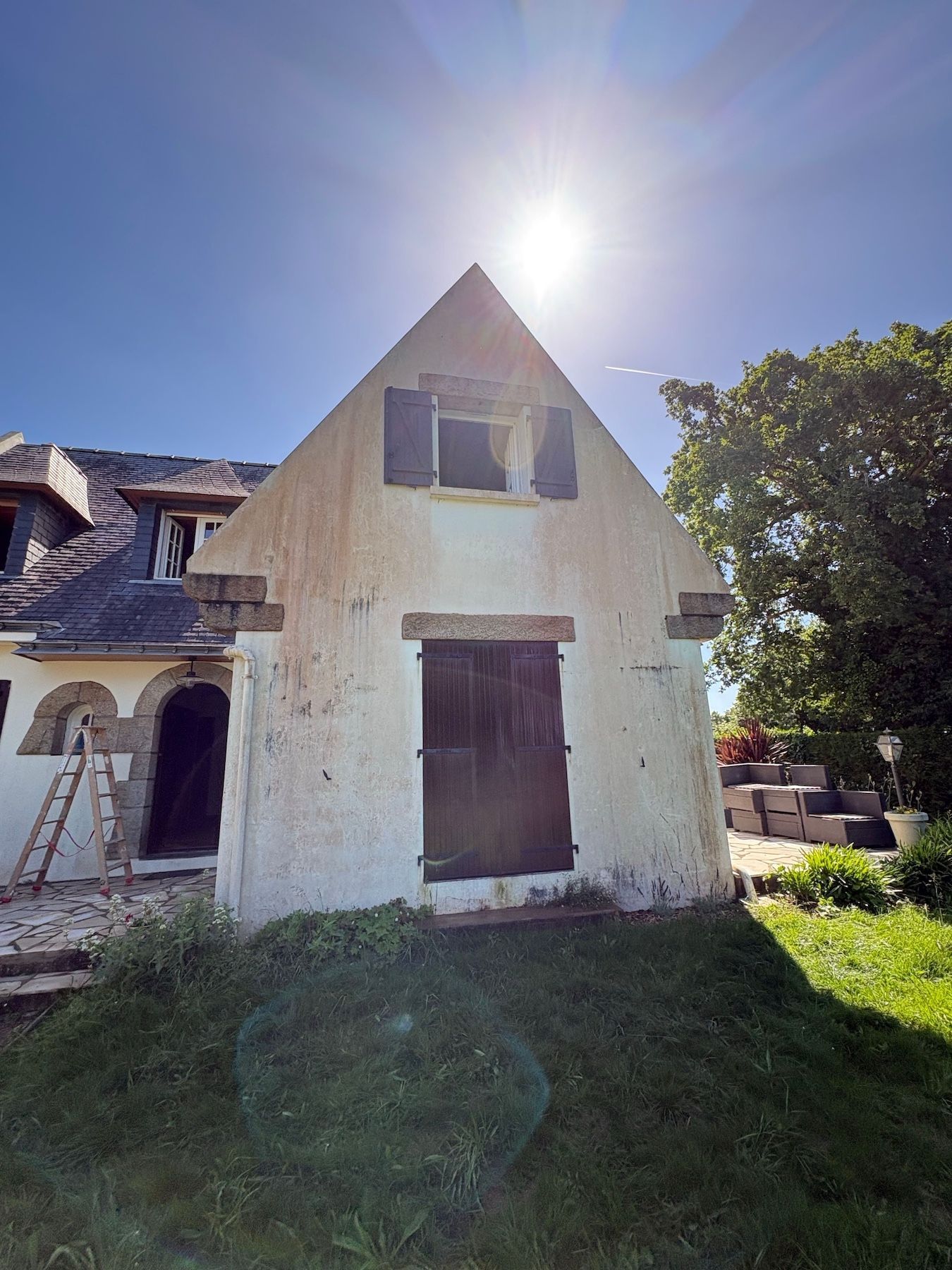 Maison blanche baignée de soleil, au toit pentu, avec porte et fenêtres en bois fermées, donnant sur une cour gazonnée.