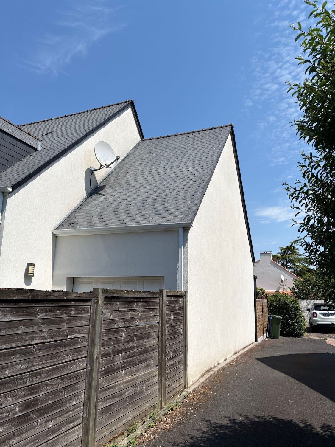 Mur latéral d'une maison blanche et allée clôturée sous un ciel bleu, au bout d'une ruelle étroite.