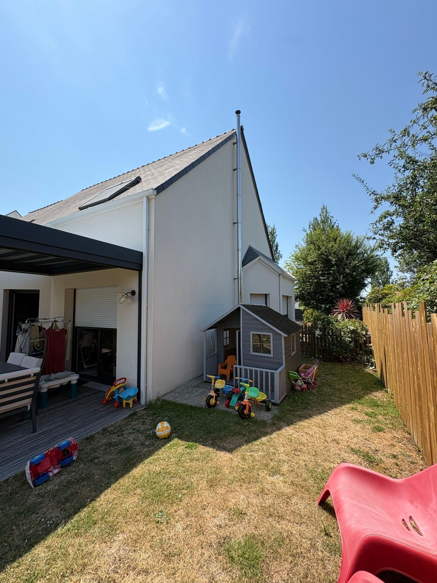 Cour arrière avec une cabane de jeux, une terrasse, une pelouse et un toboggan rouge sous un ciel bleu ensoleillé