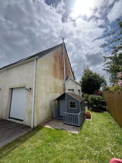 Jardin avec une petite cabane de jeux grise à côté d'une maison, terrasse en bois, pelouse et ciel nuageux.