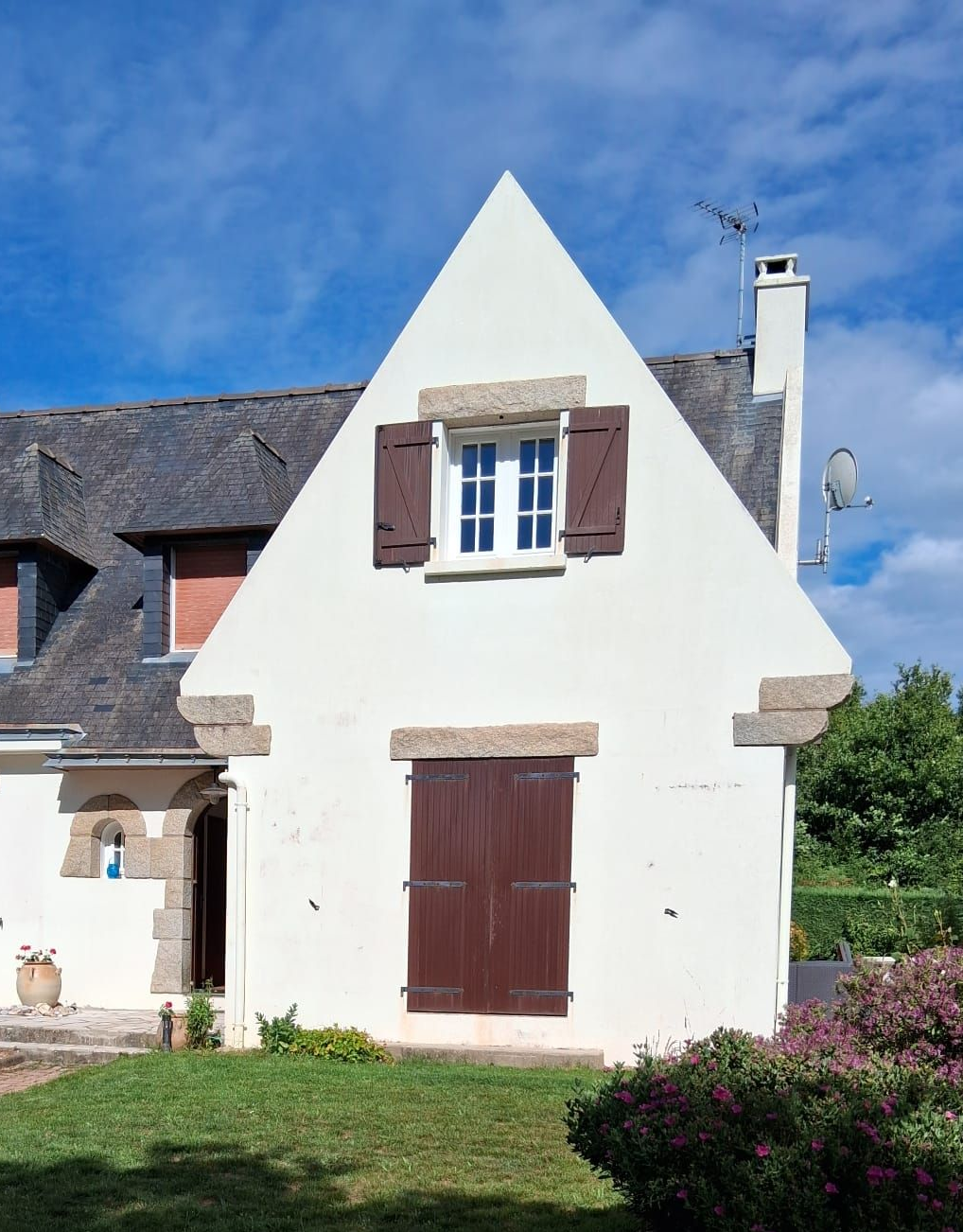 Maisonnette blanche au toit à pignon pentu, volets et porte marron, sous un ciel bleu.