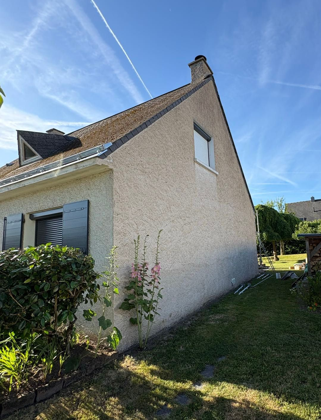 Vue latérale d'une maison en stuc crème avec un toit de tuiles, des volets et un étroit chemin herbeux à côté.