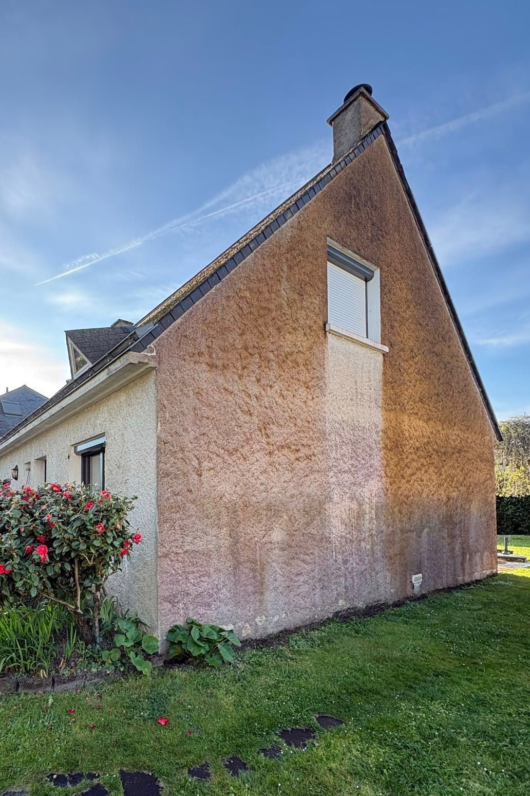 Vue latérale d'une maison en briques beiges avec un toit pentu, une fenêtre blanche et une cour herbeuse sous un ciel bleu.