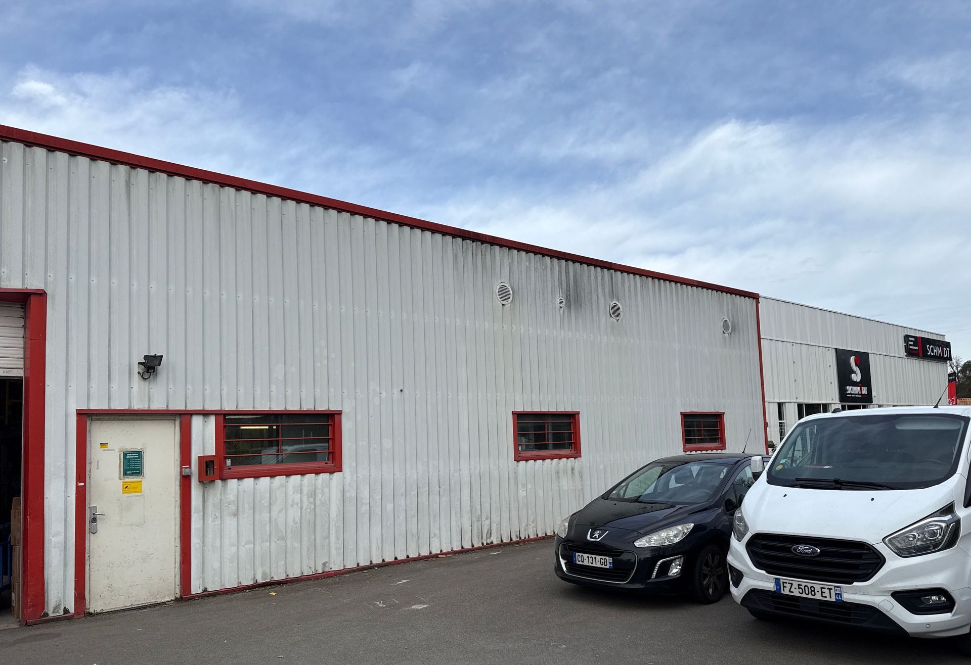 Bâtiment industriel blanc avec des bordures rouges et deux voitures garées sous un ciel nuageux