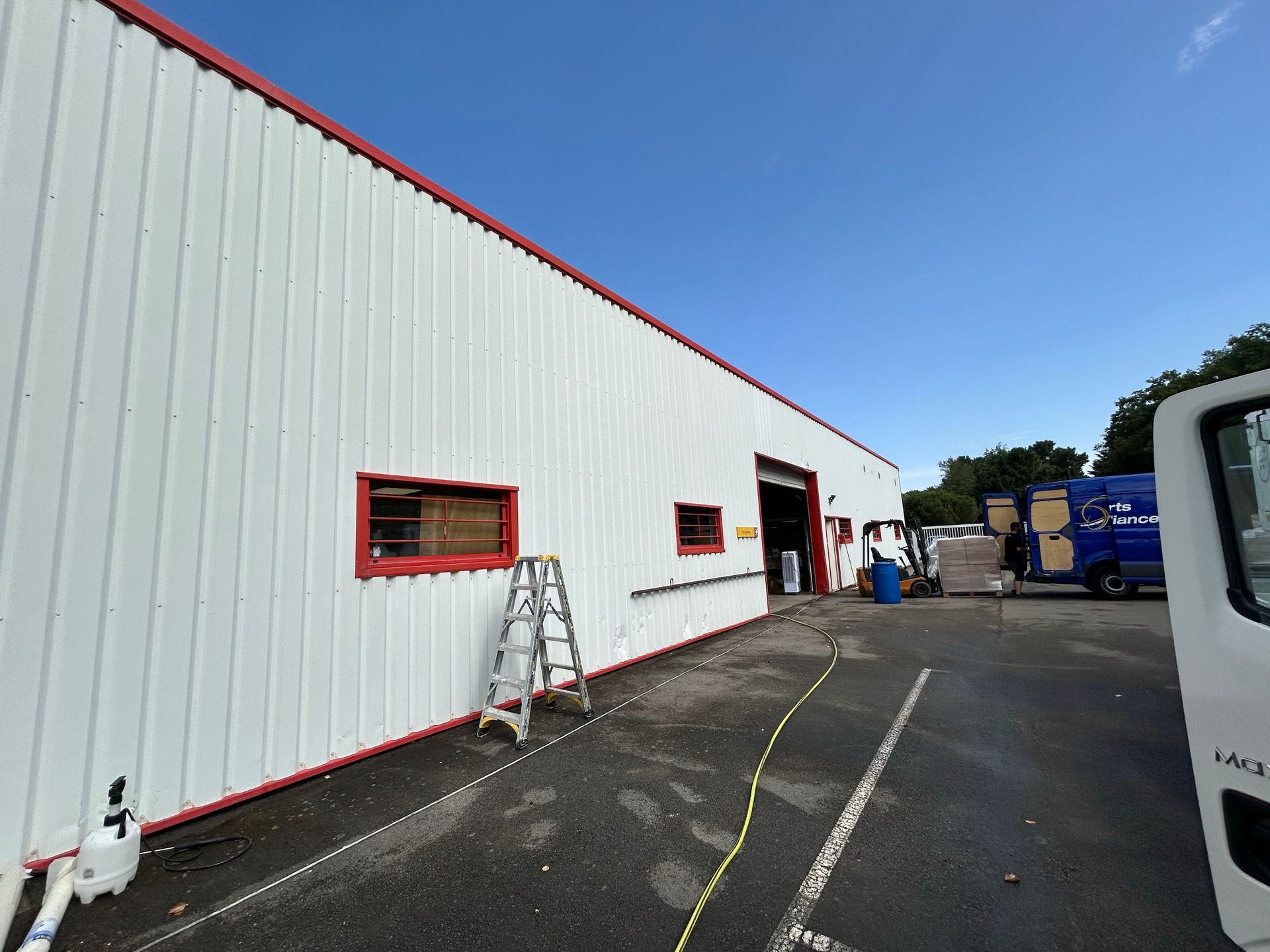 Bâtiment industriel blanc avec des finitions rouges, une échelle près d'une fenêtre et un parking à côté de camions bleus.