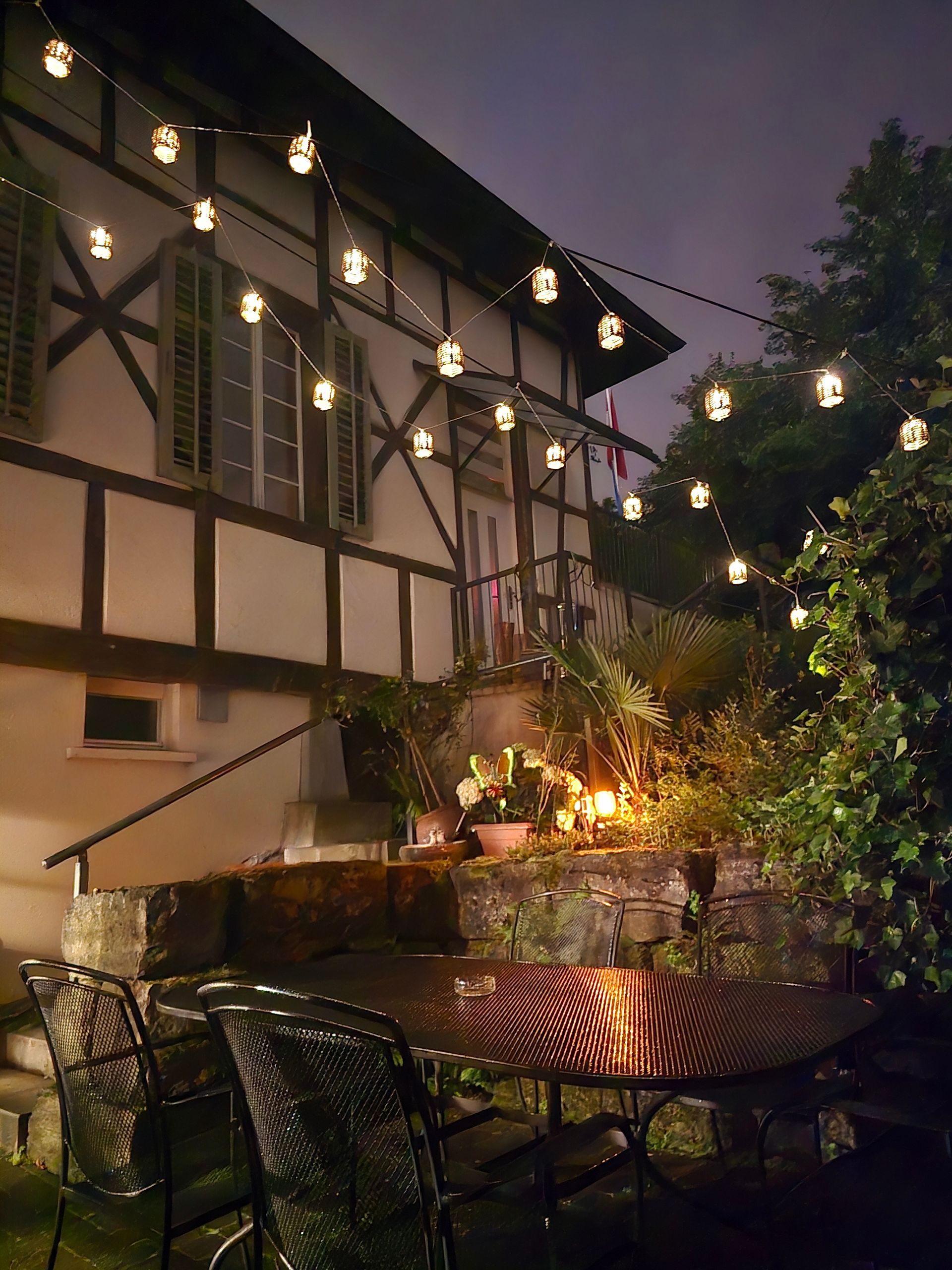 Nachtansicht eines Gebäudes mit Lichterketten über einer Terrasse. Stühle und Tisch stehen im Vordergrund.