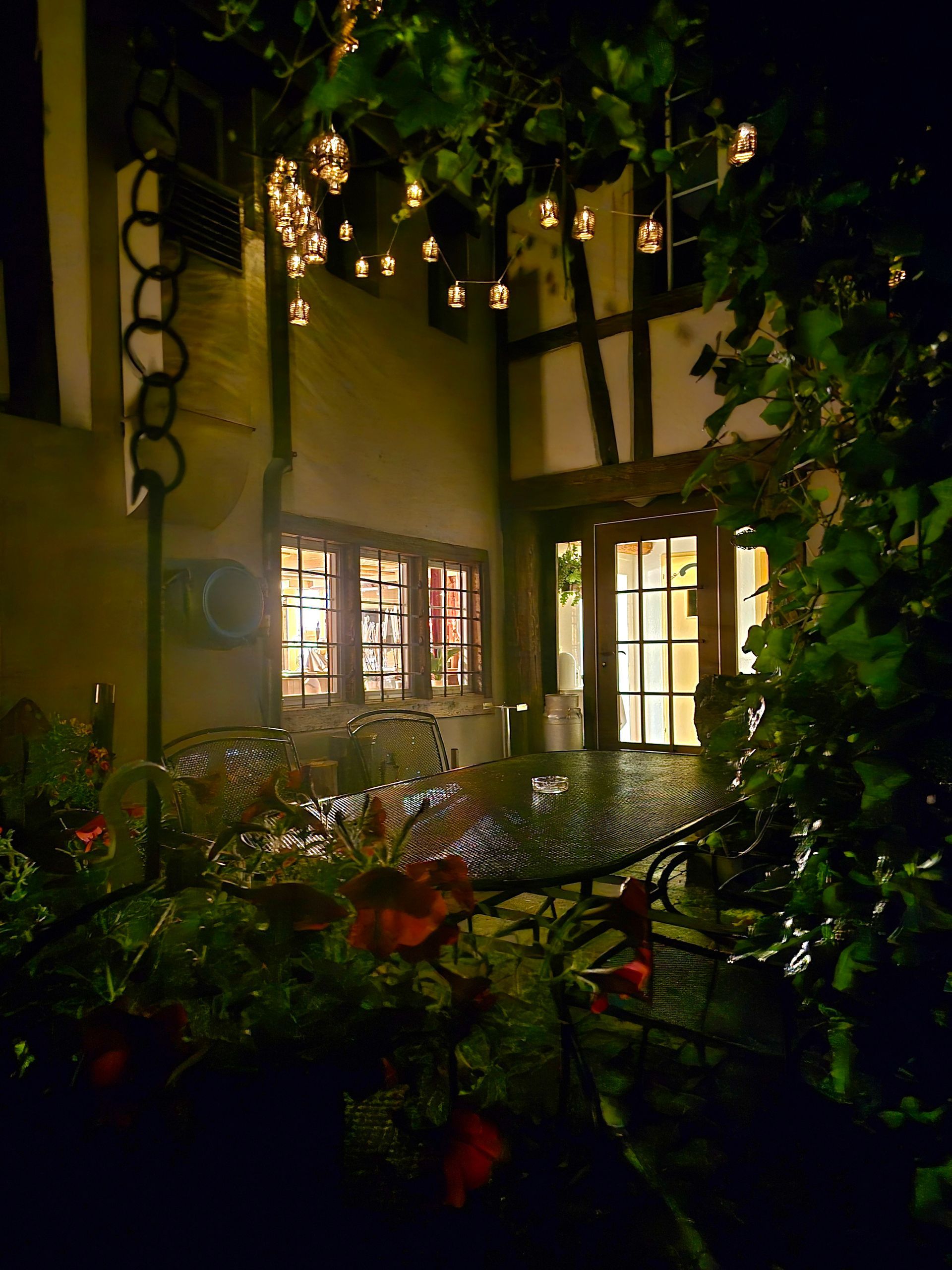 Nachtszene einer Außenterrasse mit Hängelampen, Tisch und offener Tür zu einem Gebäude.