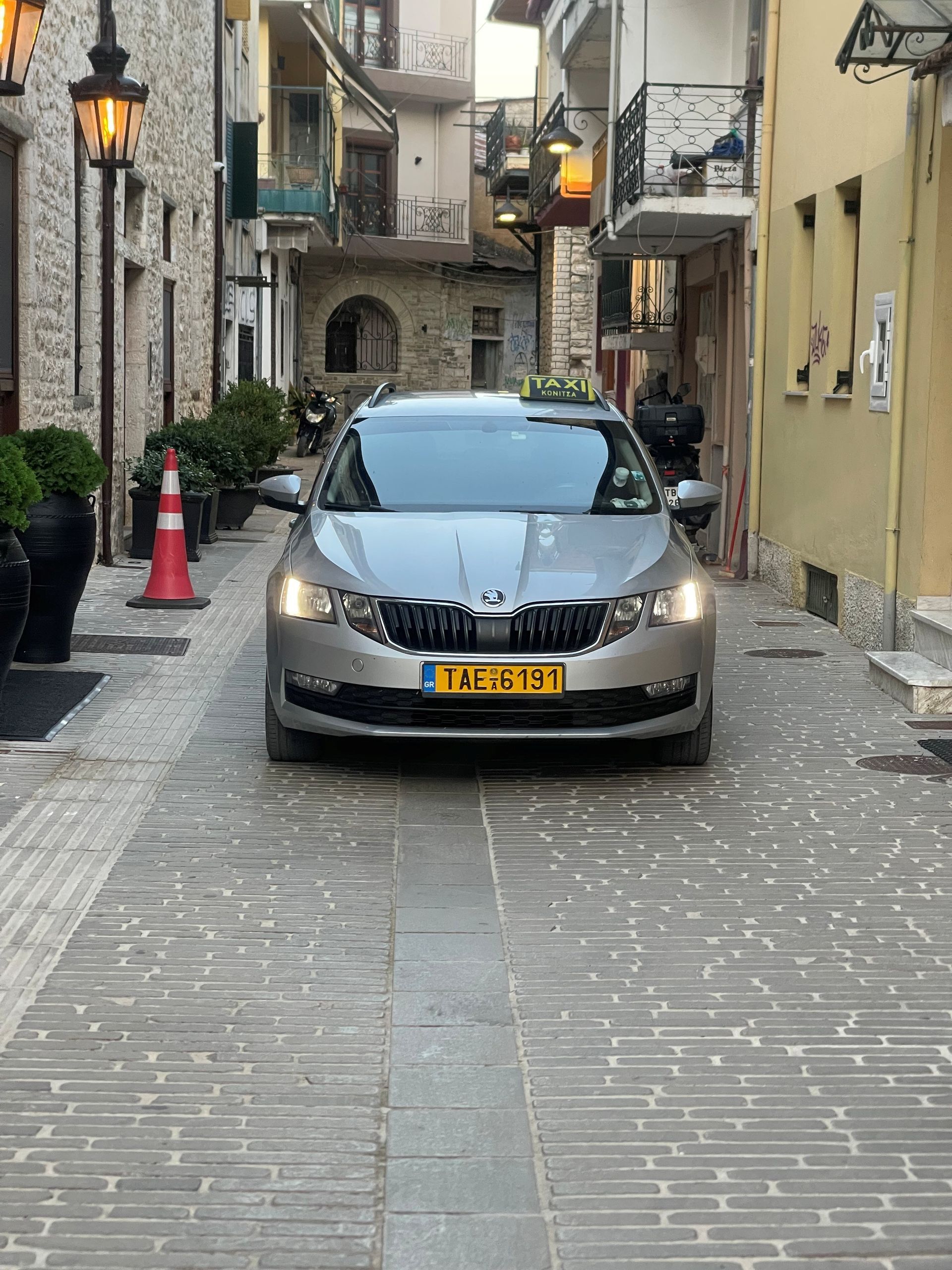 Ασημένιο αυτοκίνητο σε έναν στενό λιθόστρωτο δρόμο ανάμεσα σε παλιά πέτρινα κτίρια και ζεστά φανάρια