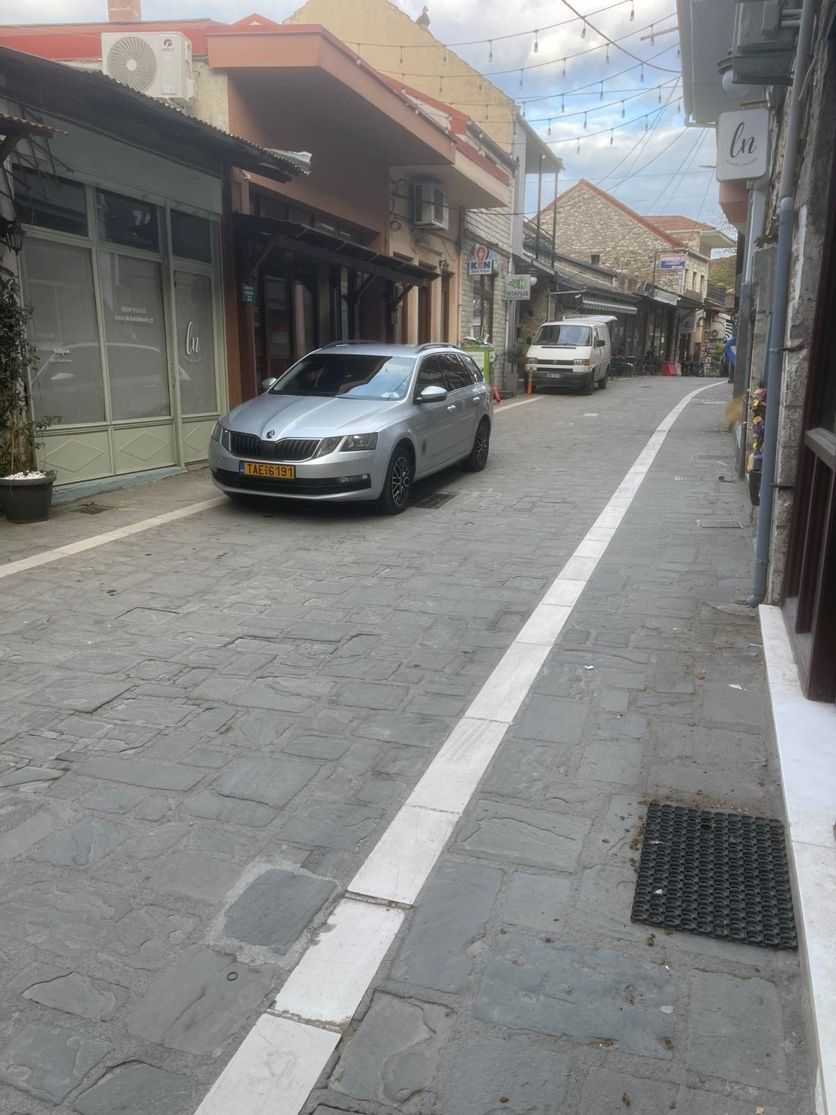 Στενός πλακόστρωτος δρόμος με παρκαρισμένα αυτοκίνητα και καταστήματα και στις δύο πλευρές, όπως φαίνεται από πεζοδρόμιο.