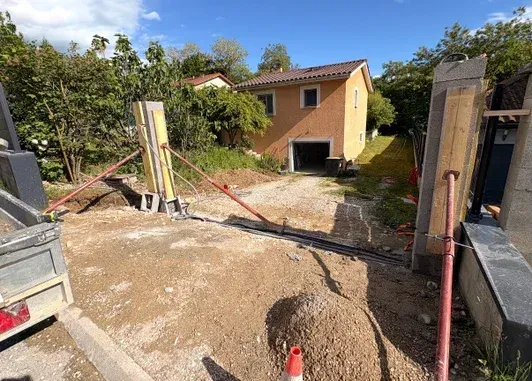 Chantier de construction avec une petite maison jaune, une allée de terre et des piliers de portail en béton.