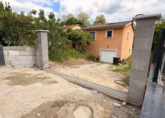 Portail d'allée partiellement construit et maison sur un terrain en terre battue, avec des piliers en pierre.