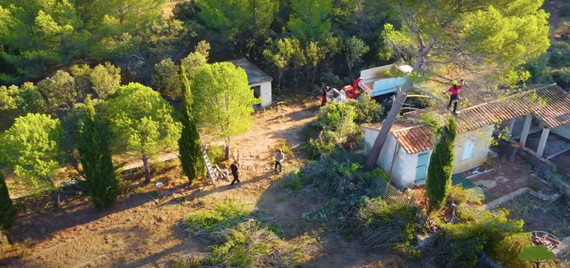 Vue aérienne d'un jardin boisé avec un petit bâtiment blanc, un tracteur rouge et une personne près des arbres et des clôtures.