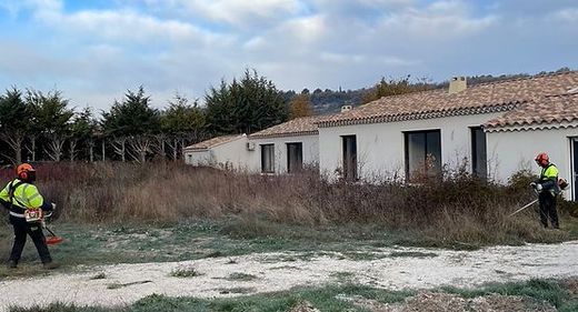 Deux ouvriers munis de débroussailleuses dégagent les broussailles envahissantes à côté d'un long bâtiment blanc.
