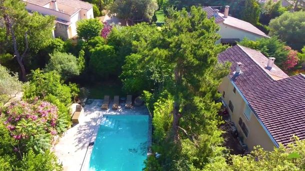 Vue aérienne d'une piscine dans un jardin entouré d'une végétation luxuriante et de maisons aux toits de tuiles.