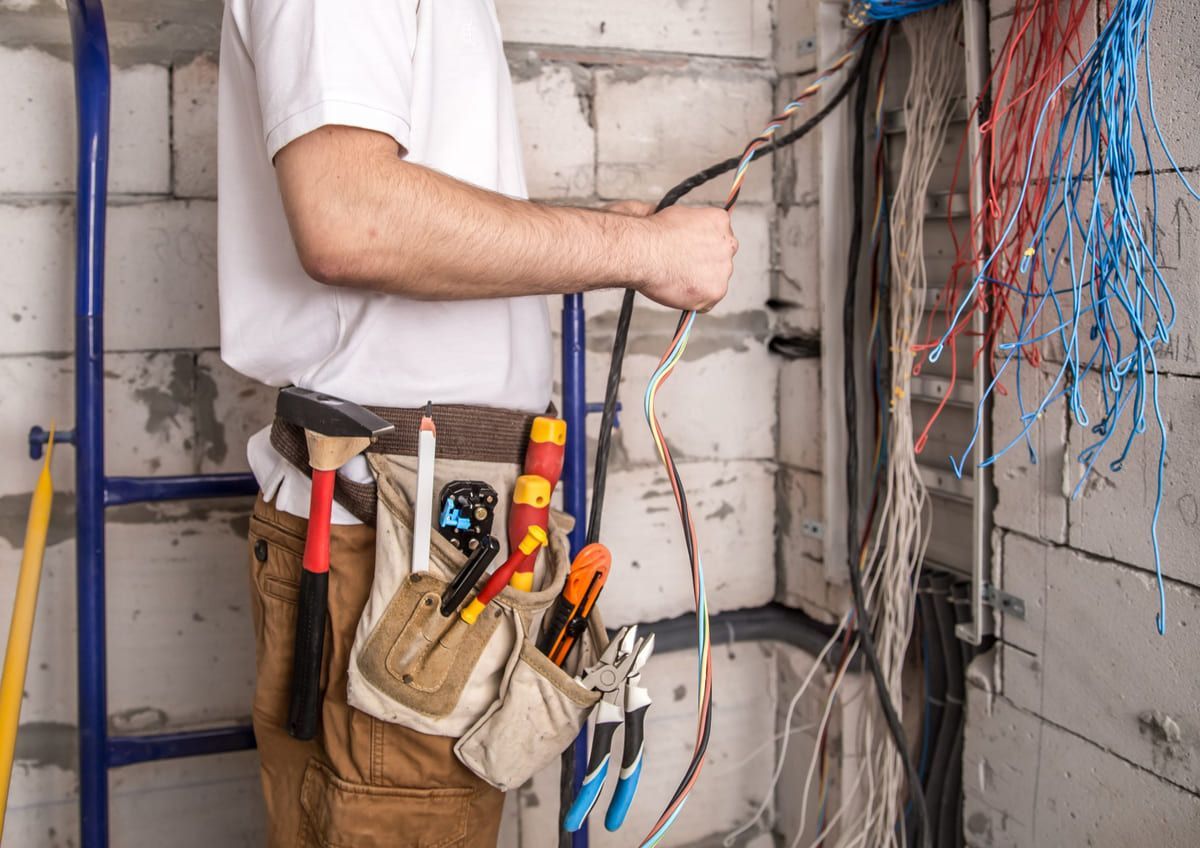 Electricista sosteniendo herramientas junto al cableado expuesto en una pared durante un trabajo eléctrico.