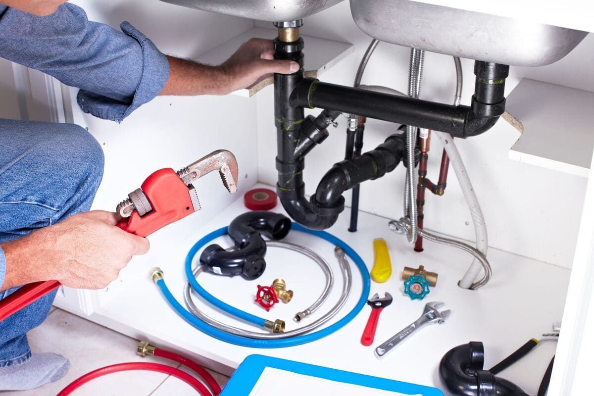 Fontanero reparando las tuberías del lavabo debajo de un mueble de baño con herramientas y accesorios cerca.