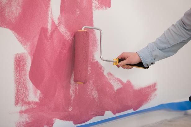 Persona pintando una pared de rosa con un rodillo, con cinta azul a lo largo del borde inferior.