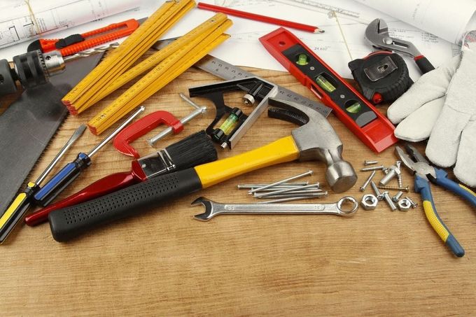 Varias herramientas manuales y ferretería sobre un banco de trabajo de madera, incluyendo martillo, llave inglesa, cinta métrica y guantes.