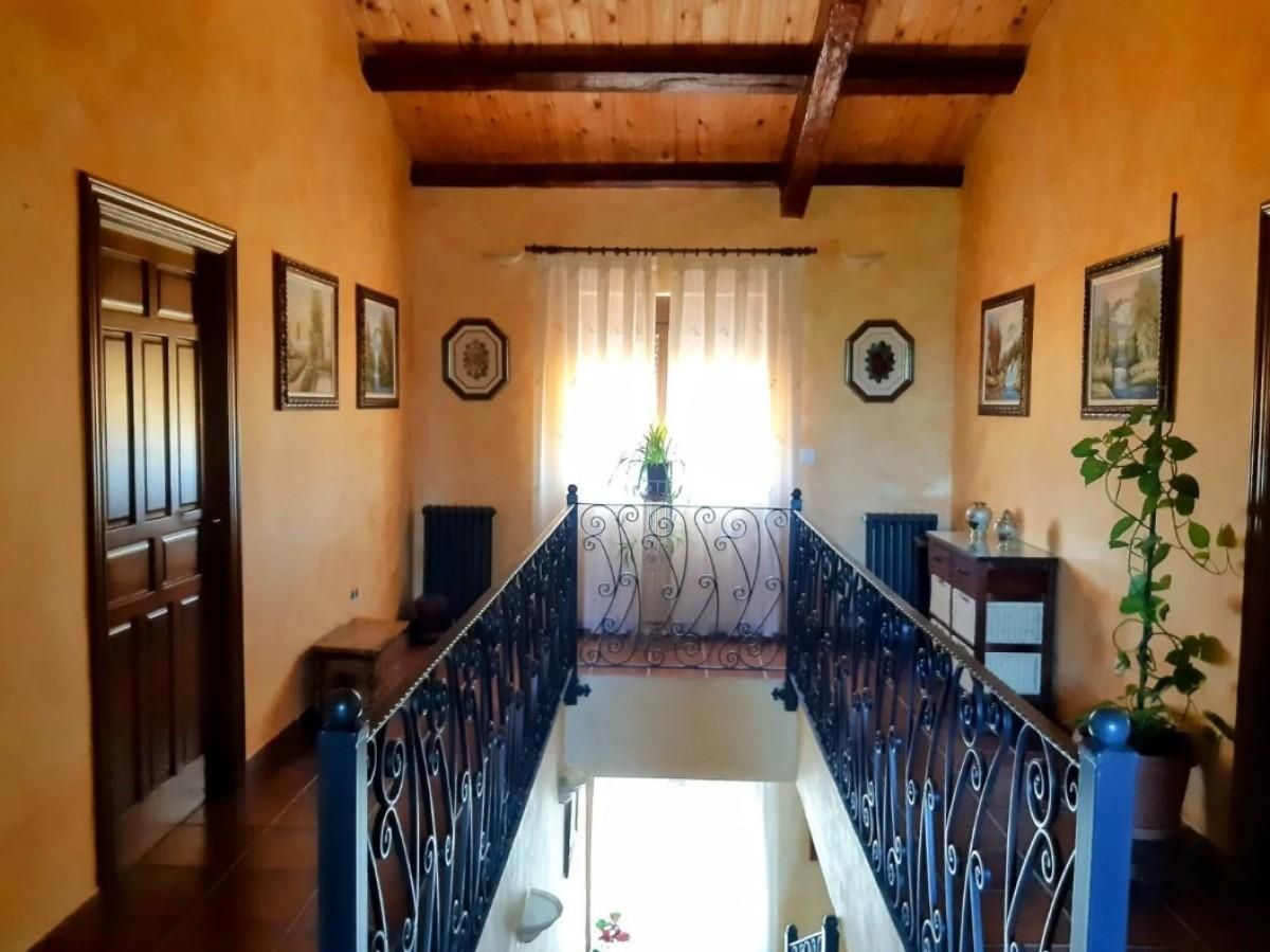 Pasillo de tonos cálidos con barandilla de hierro forjado, vigas de madera, obras de arte en la pared y una luminosa ventana al final.