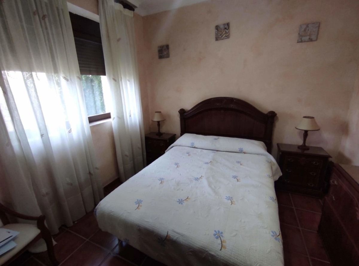 Habitación pequeña con cama hecha, cabecero de madera oscura, mesita de noche y cortinas transparentes junto a la ventana.