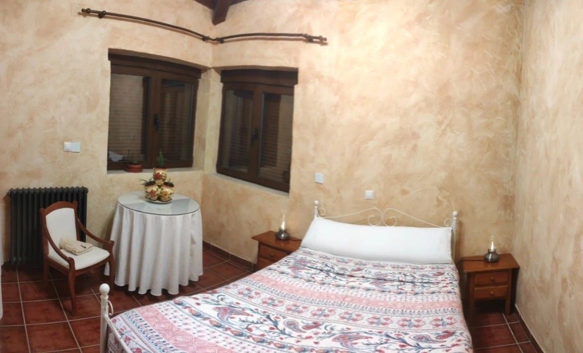 Habitación acogedora con cama blanca, colcha estampada, ventanas de madera oscura y una pequeña mesa redonda con silla.