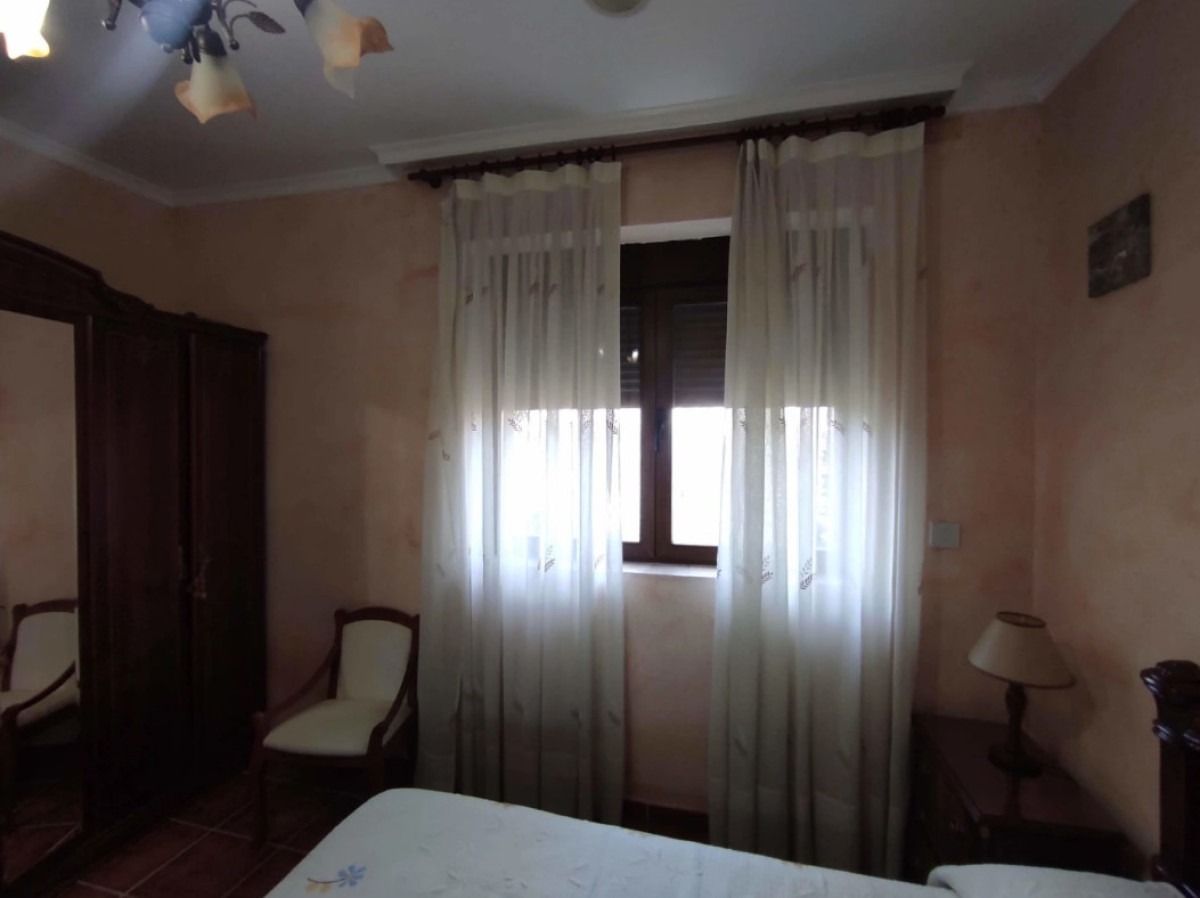 Habitación con poca luz, con ventana, cortinas, dos sillas, un armario y una cama parcialmente visible.