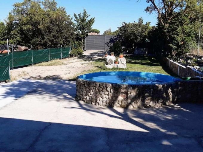 Patio trasero con una cama elástica redonda azul, base de piedra, árboles y sillas de patio bajo la luz del sol.